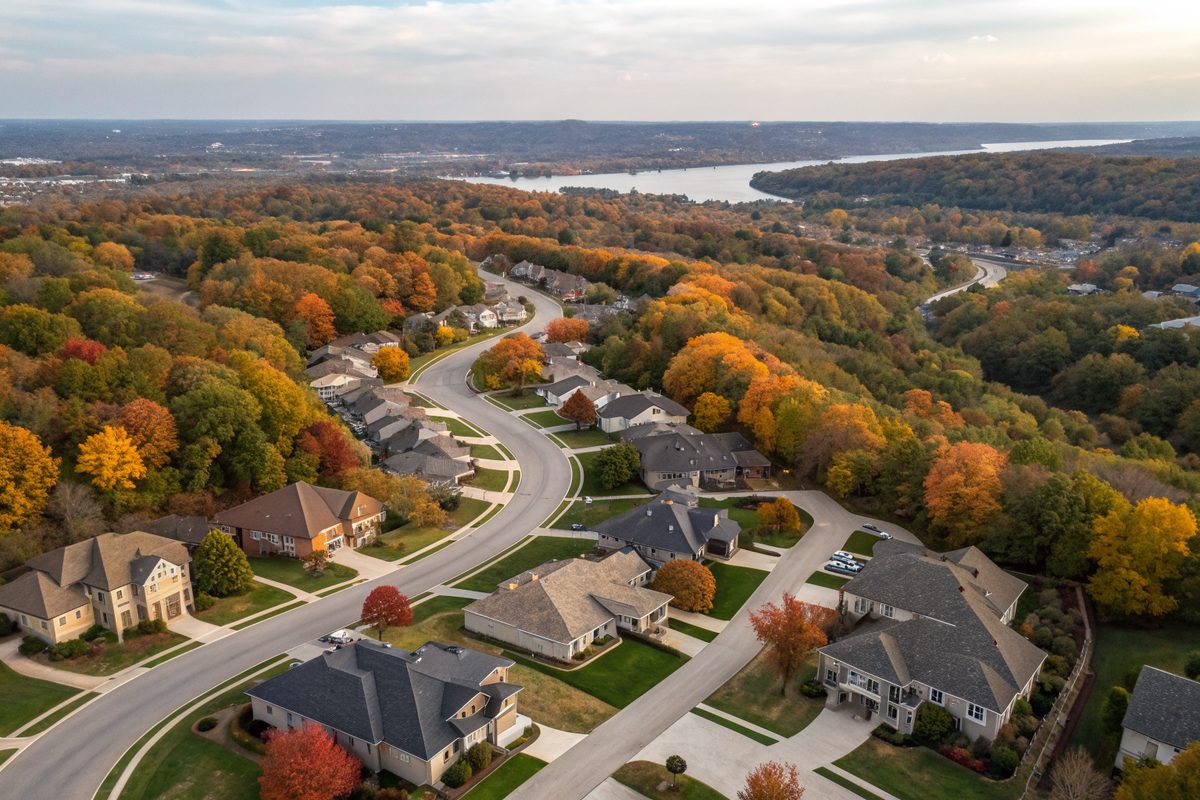 Aerial view of shingle-roofed homes on curving streets among wooded hills in fall color, with a river in the distance