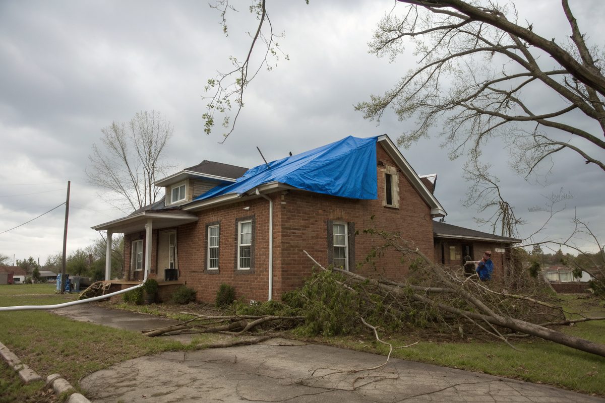 A brick house with a blue tarp over part of its roof and broken branches on the lawn