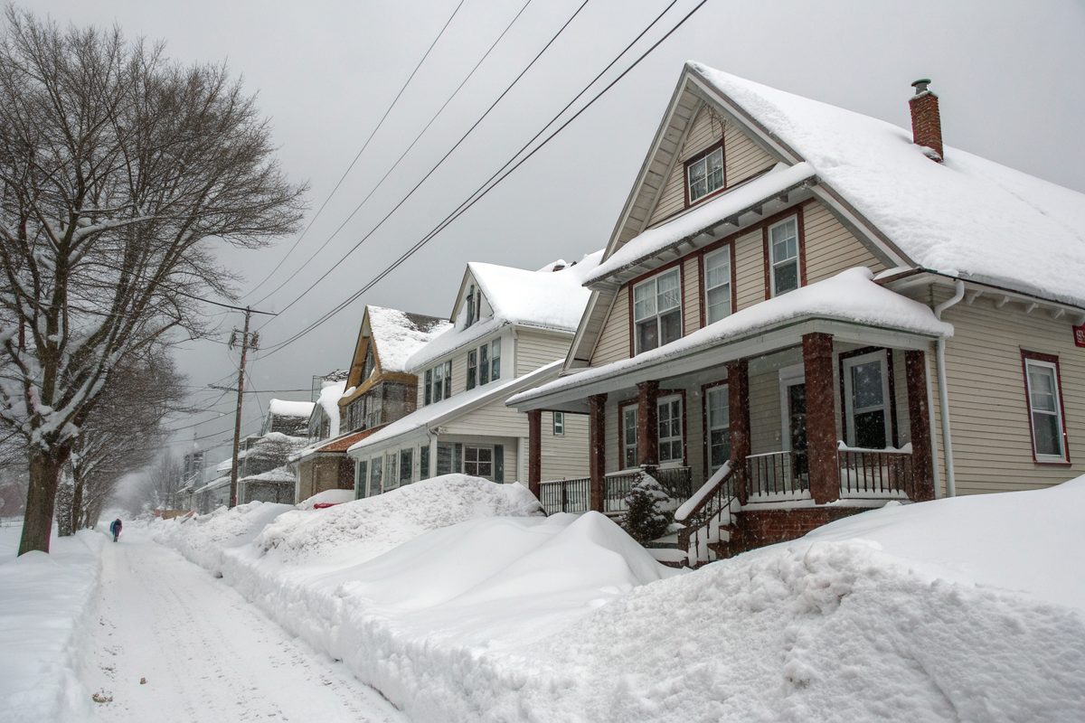 Deep snow piled on the roofs and sidewalks of a street of older two-story houses